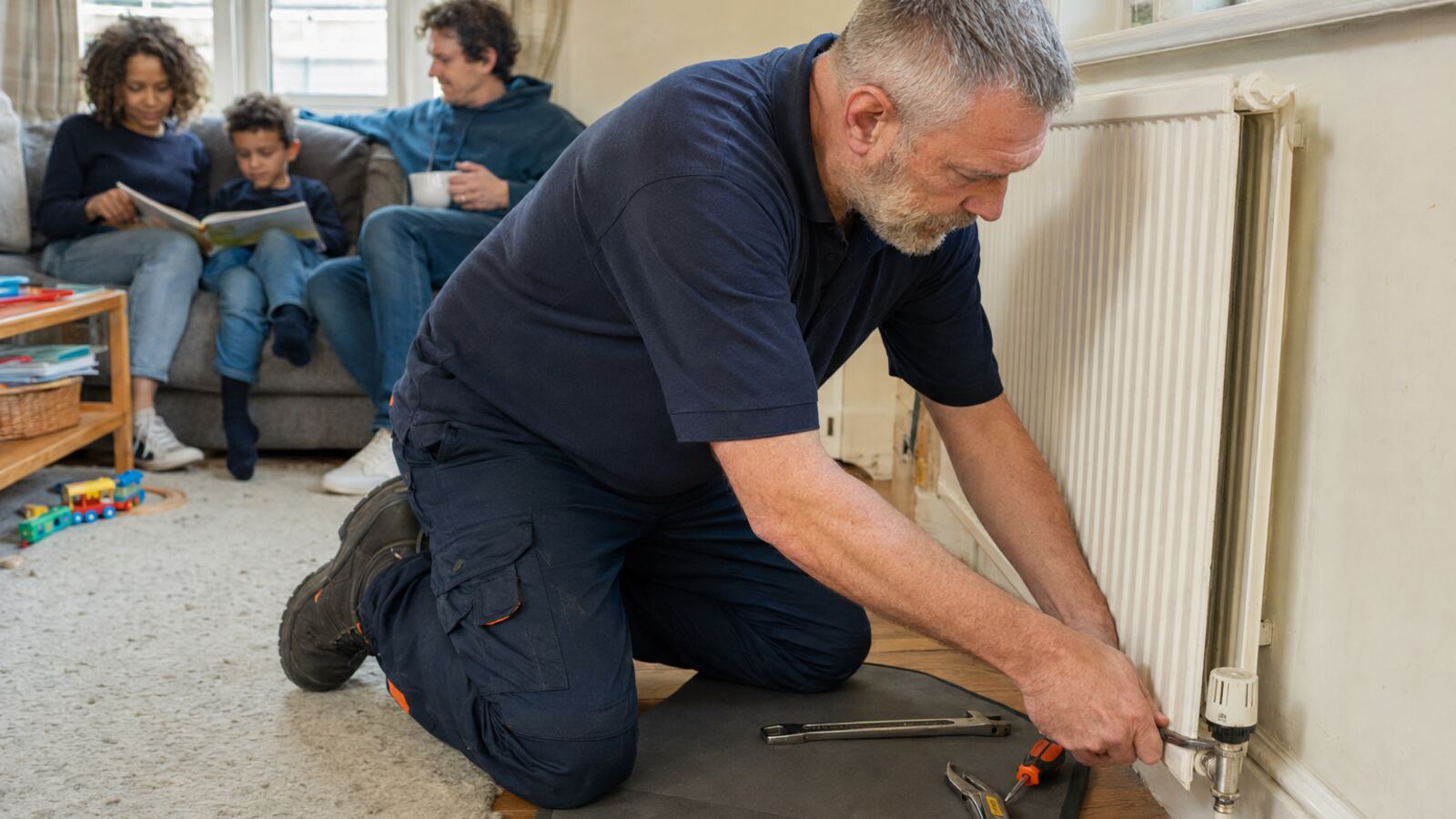 Heating engineer making a final radiator adjustment while a family relaxes nearby